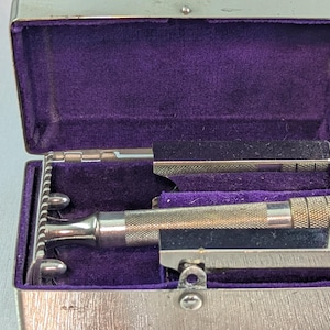 Könnte beinhalten: Ein silberner Sicherheitsrasierer mit einem mit violettem Samt ausgekleideten Etui. Der Rasierer hat eine verchromte Oberfläche und eine Sicherheitsleiste mit einer Klinge.