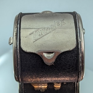 May include: Close-up of a vintage Twinplex razor case. The case is black leather with a silver metal top. The word "Twinplex" is embossed on the top. The case has a clasp and is likely from the early 20th century.