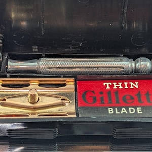 Puede incluir: Juego de afeitadora Gillette vintage en un estuche negro. El juego incluye un mango de afeitadora plateado, un cabezal de afeitadora dorado y una caja roja y blanca de cartuchos "Thin Gillette Blade". El estuche está abierto, mostrando el contenido.