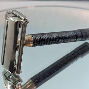 Puede incluir: Una maquinilla de afeitar de seguridad vintage con cabezal plateado y mango negro texturizado. La maquinilla se muestra sobre una superficie reflectante, con su reflejo debajo. El mango tiene un patrón moleteado para agarre.