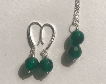 Puede incluir: Un conjunto de pendientes y collar de piedras preciosas verdes. Los pendientes son de plata con una piedra preciosa verde que cuelga de cada uno. El collar es una cadena de plata con dos piedras preciosas verdes.