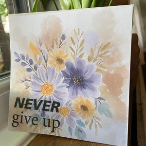 May include: Square canvas art featuring a watercolor floral design in shades of purple, yellow, and gold. The words "NEVER give up" are printed in black. The artwork is displayed near a window with a plant.