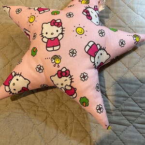 Pode incluir: Almofada rosa em forma de estrela com um padrão repetido de Hello Kitty, flores, abelhas e sóis sorridentes. A almofada é feita de tecido macio e tem uma etiqueta. Um item decorativo para o quarto de uma criança.