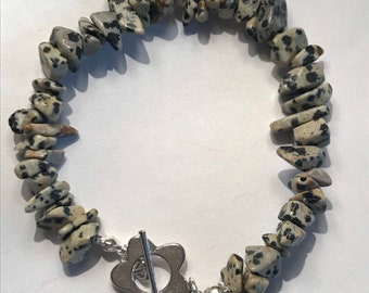 May include: A bracelet made of small, irregular, black and white dalmatian jasper stones. The bracelet has a silver clasp in the shape of a flower.