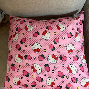 Pode incluir: Almofada rosa com um padrão repetido de rostos de gatos de desenho animado brancos com laços vermelhos e morangos rosa. O fundo é rosa claro com pequenas bolinhas brancas. A almofada está em uma superfície cinza.