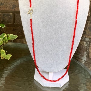 May include: A red beaded necklace with a silver toggle clasp. The necklace is displayed on a white mannequin.