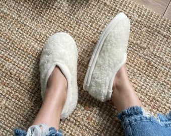 Può includere: Un paio di pantofole bianche e soffici con un design semplice. Le pantofole sono indossate da una persona che indossa jeans blu e sono sedute su un tappeto intrecciato.