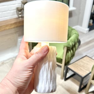 Puede incluir: Una lámpara de mesa blanca con una base blanca con patrón geométrico y una pantalla de tela blanca. La lámpara está siendo sostenida en la mano de una persona.