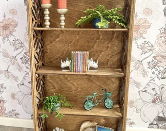 May include: A wooden bookshelf with four shelves, featuring decorative items such as candles, plants, books, a bicycle, coral, and a framed photo. The bookshelf has a geometric design on the sides.