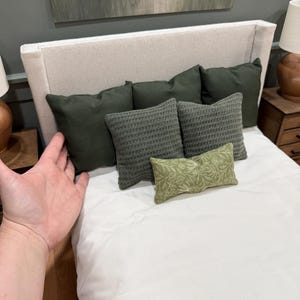 May include: A miniature bedroom scene featuring a cream headboard and white bedding. The bed is dressed with several green pillows in different textures and patterns. The scene is set against a neutral wall, creating a cosy and inviting aesthetic.