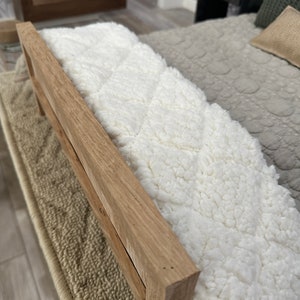 May include: A light-coloured wooden bed frame with a fluffy white blanket. The blanket has a quilted pattern. The bed frame is rectangular and has a natural wood grain. The bed is on a beige rug.