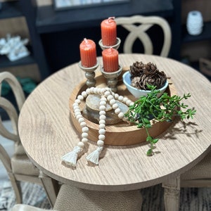 May include: A wooden tray with a white beaded garland, three orange candles in silver candle holders, a small bowl of pine cones, and a potted green plant.