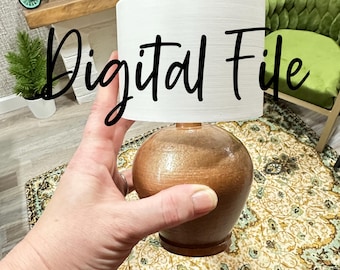 Può includere: Una lampada in miniatura con base marrone e paralume bianco. Il testo "Digital File" è scritto sul paralume. La lampada è tenuta in mano e lo sfondo comprende una sedia verde e un tappeto decorativo.