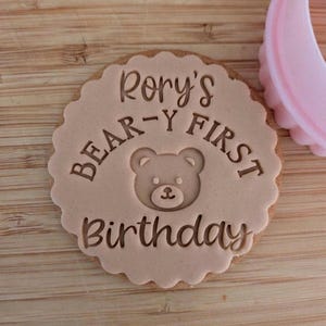 May include: A round, light brown biscuit with a scalloped edge featuring an embossed teddy bear face in the centre. The text "Rory's BEAR-Y FIRST Birthday" is imprinted around the bear. A pink plastic biscuit cutter sits to the right.