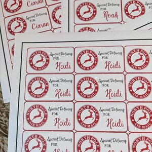 Puede incluir: Una hoja de papel blanco con etiquetas de regalo de Navidad rojas y blancas. Las etiquetas muestran un reno rojo con las palabras "North Pole Air Mail" y "Special Delivery" impresas en ellas. Las etiquetas est&aacute;n personalizadas con los nombres "Ciaran", "Noah" y "Heidi".