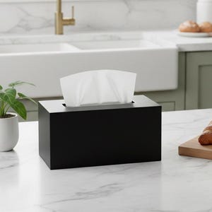 May include: A black rectangular tissue box with white tissues peeking out. The box sits on a white marble countertop in a kitchen setting. A small potted plant and a mug are also on the counter.