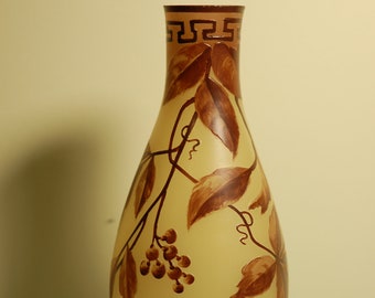 May include: A yellow glass vase with a brown and white floral design. The vase has a narrow neck and a wide base. The floral design features leaves and berries.