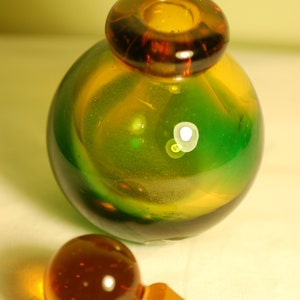 May include: A green and amber glass bottle with a round body and a separate amber glass stopper. The bottle has a swirl pattern and a textured surface.