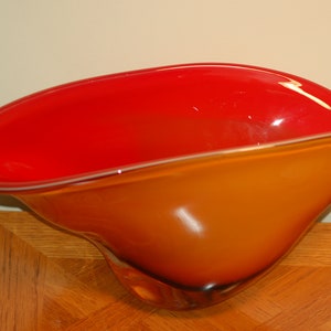May include: A large, red and orange glass bowl with a wavy, organic shape. The bowl is made of clear glass with a red interior and an orange exterior.