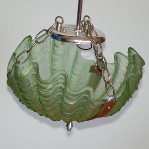 Puede incluir: Una lámpara colgante de vidrio verde con un diseño ondulado en forma de concha. La luz está suspendida por una cadena plateada y una cubierta de metal. La lámpara tiene una textura decorativa y un pequeño remate plateado.