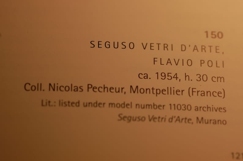 May include: Close-up of a beige label with text describing a Seguso Vetri dArte glass piece by Flavio Poli, circa 1954.  The label notes the height (30 cm), collection (Nicolas Pecheur, Montpellier, France), and archival listing (model number 11030).  The labels text is in a serif typeface, and the background is a muted beige color.  The number 150 is prominently displayed at the top.