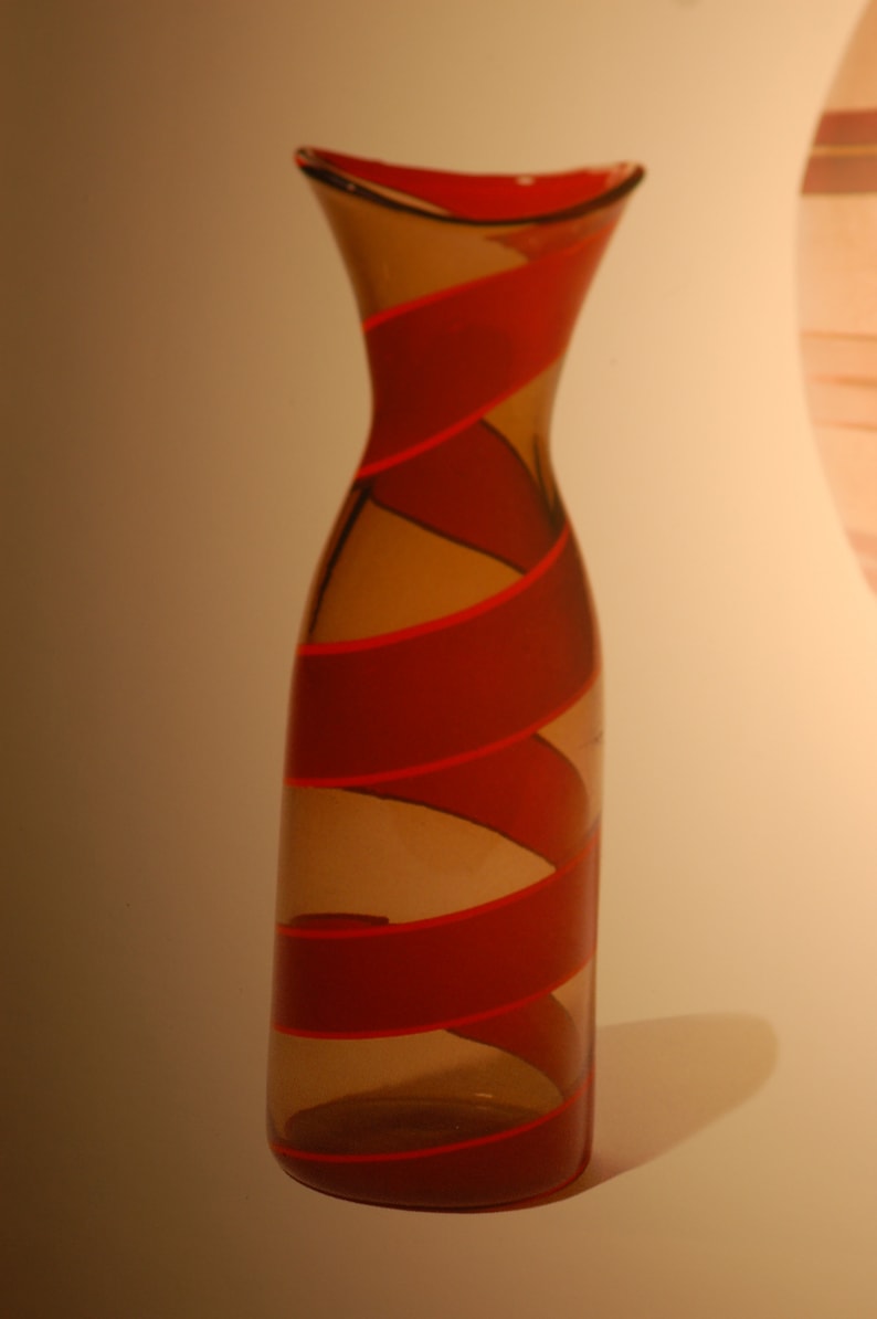 May include: A decorative glass vase with a unique design. The vase is amber-brown with red spiral stripes. The glass is transparent, allowing the color to show through. The vase has a slender, curved shape, tapering slightly at the base and neck. Its a stylish home decor piece, perfect for adding a touch of modern elegance to any room. The red and amber color scheme is eye-catching and versatile, complementing various interior styles.