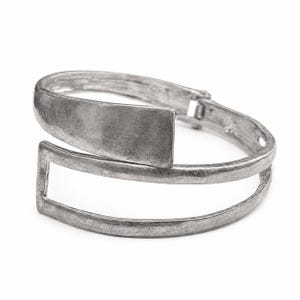 May include: A silver-toned metal cuff bracelet with a rectangular plate and a double-band design. The bracelet has a hinged clasp and a brushed finish, creating a vintage look. The bracelet is shown on a white background.