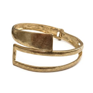 May include: Gold-toned metal bracelet with a rectangular plate and open rectangular design. The bracelet has a hinged clasp for easy wear and measures approximately 2 inches in diameter.