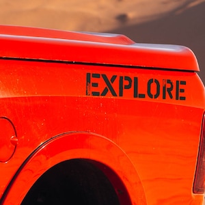 Può includere: Un pick-up rosso con un adesivo nero "EXPLORE" sul lato. Il pick-up è parcheggiato in un ambiente desertico.