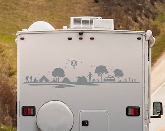 Op de afbeelding: Een grijze camper met een sticker van een kampeerscène op de achterkant. De sticker toont een heteluchtballon, bomen, tenten en mensen die aan het kamperen zijn.