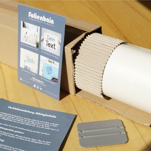 Może przedstawiać: Biały rulon folii matowej do okien w kartonowym pudełku z szarym rakle. Pudełko jest oznaczone "Folienhain" i zawiera instrukcje dotyczące nakładania folii.
