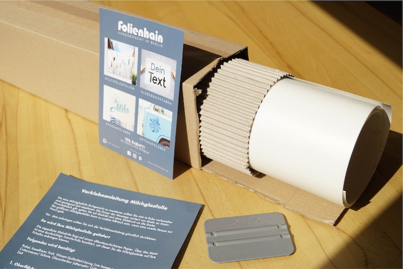Może przedstawiać: Biały rulon folii matowej do okien w kartonowym pudełku z szarym rakle. Pudełko jest oznaczone "Folienhain" i zawiera instrukcje dotyczące nakładania folii.