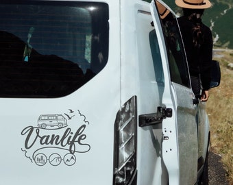 May include: A white van with a black and white sticker on the back that says "Vanlife" with a cartoon van, birds, and three icons representing trees, waves, and mountains.