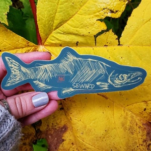 May include: A blue and white sticker of a salmon with the text "SEWARD" and "AK" on it. The sticker is on a yellow leaf.