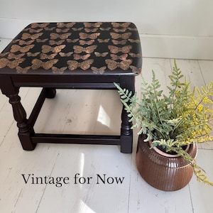May include: A dark brown wooden footstool with a black top decorated with copper-coloured butterfly patterns. The stool has turned legs and a rectangular seat. The text "Vintage for Now" is at the bottom of the image.