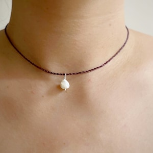 May include: A delicate choker necklace featuring a dark brown cord. A small, white, pear-shaped pendant hangs from the center. The necklace is worn around the neck, showcasing a minimalist jewelry design.