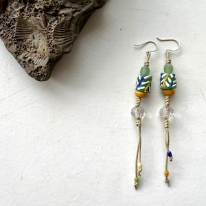 Puede incluir: Pendientes hechos a mano con ganchos de plata, con cuentas estampadas verdes, azules y blancas, cuentas de vidrio transparente y pequeños acentos coloridos. Los pendientes cuelgan de un cordón beige.