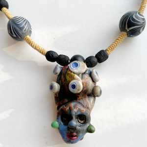 May include: A handmade necklace with a distinctive face-shaped pendant. The pendant is a mix of colours, including blue, red, green, and yellow. The necklace also has black and white beads and a beige rope.