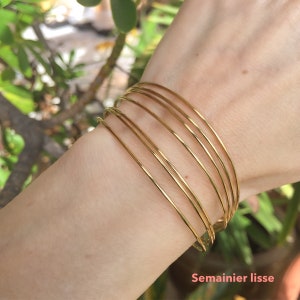 Peut inclure: Plusieurs bracelets dorés fins et lisses empilés sur un poignet. La lumière se reflète sur les bracelets. Le texte "Semainier lisse" est visible en bas à droite de l'image. Un accessoire simple et élégant.