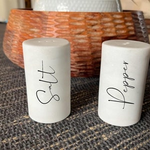 May include: Two white ceramic salt and pepper shakers with black script lettering. The shaker on the left says "Salt" and the shaker on the right says "Pepper".