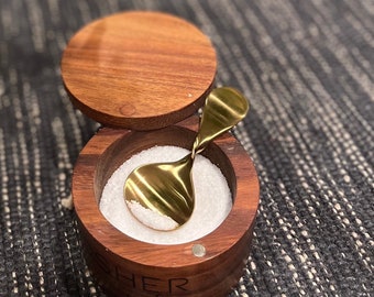 May include: A wooden salt cellar with a gold-colored spoon inside. The cellar is engraved with the words "Kosher Salt".