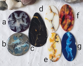 May include: Seven oval-shaped coasters with abstract designs in various colors, including black, white, blue, yellow, orange, and purple. The coasters are arranged in a semi-circle on a white surface.