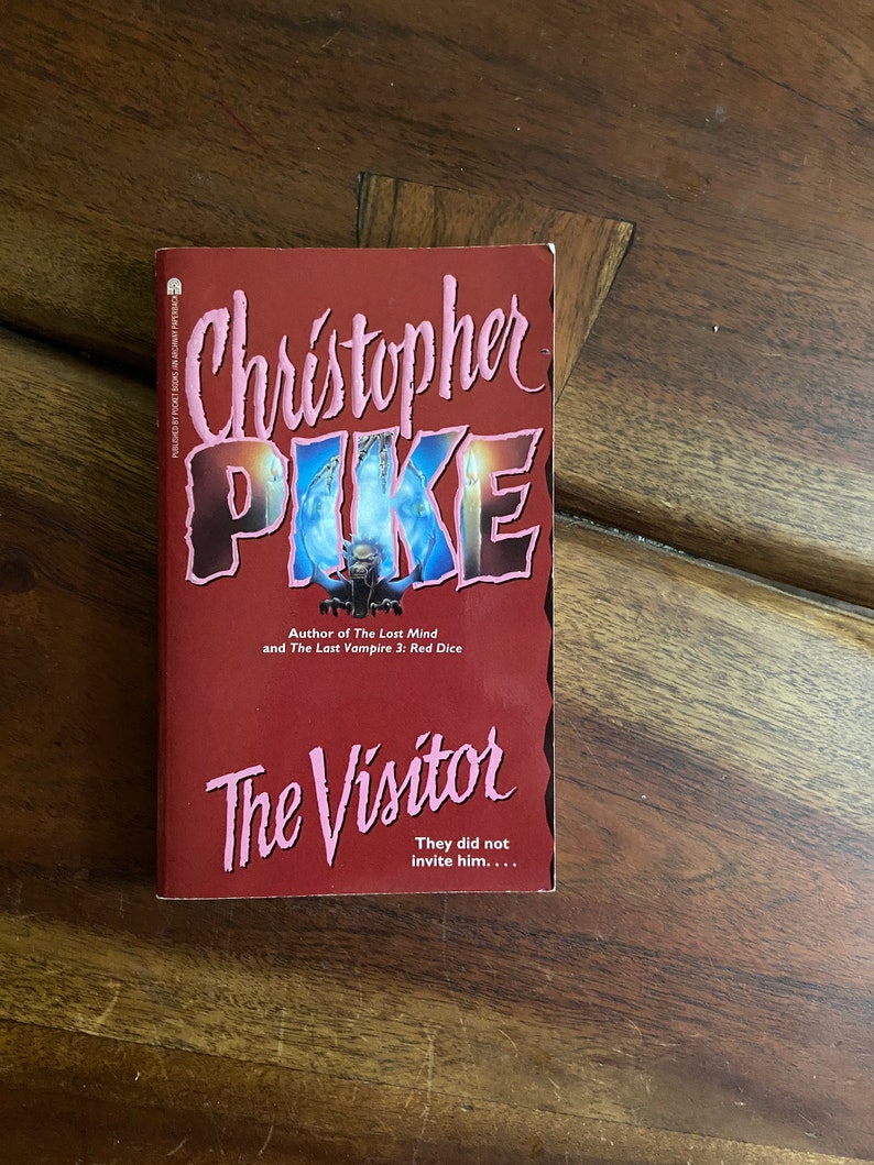 Może przedstawiać: Czerwona książka w miękkiej oprawie zatytułowana "The Visitor" autorstwa Christophera Pike'a. Okładka przedstawia postać w cieniu z błyszczącymi oczami i świecami.