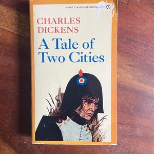 May include: A vintage paperback book titled "A Tale of Two Cities" by Charles Dickens. The cover features an illustration of a man in a black hat and coat. The book has an orange border and the title is in blue.