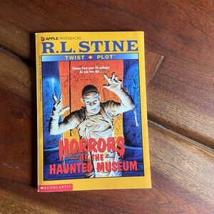 May include: A yellow paperback book with a red and blue illustration of a mummy on the cover. The book title is "Horrors of the Haunted Museum" by R.L. Stine. The book is part of the "Twist a Plot" series.