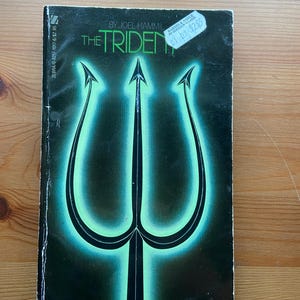 May include: A paperback book titled "The Trident" by Joel Hammil. The cover features a glowing green trident design against a dark background. The book has a price sticker on the top right corner.