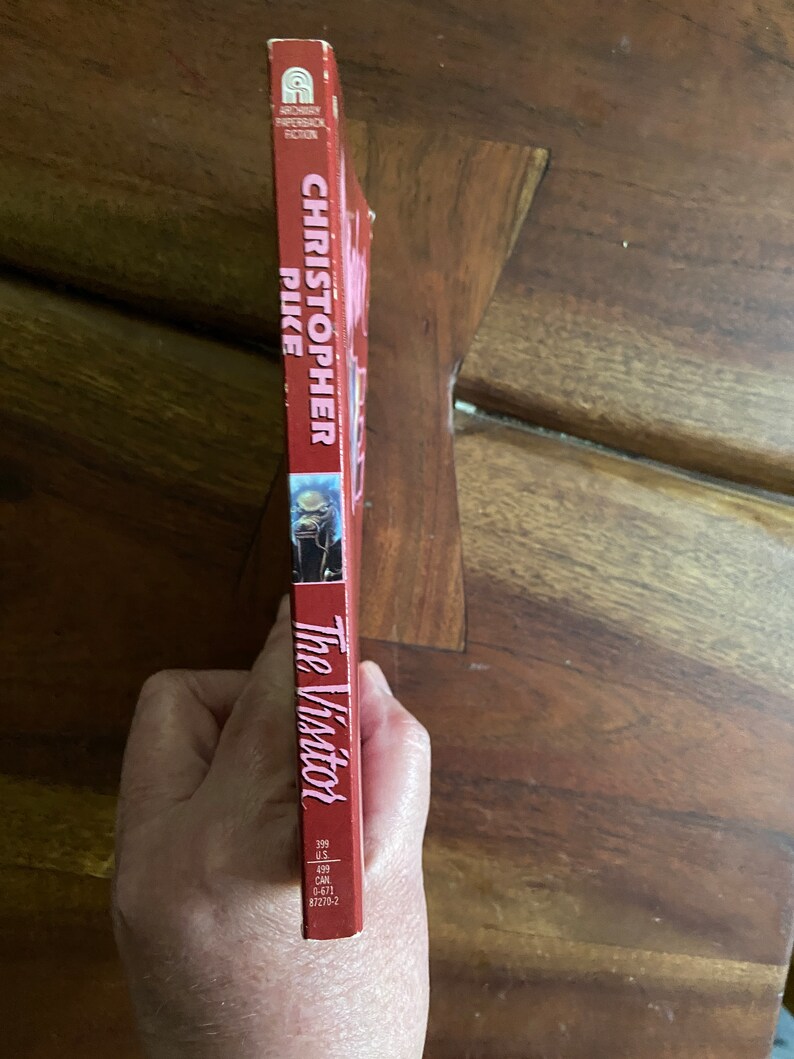 Może przedstawiać: Czerwona książka w miękkiej oprawie zatytułowana "The Visitor" autorstwa Christophera Pike'a. Książka jest częścią serii Archway Paperback Fiction.