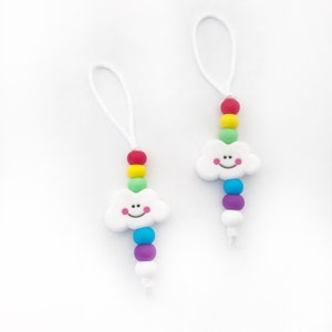Könnte beinhalten: Zwei weiße wolkenförmige Silikon-Anhänger mit lächelnden Gesichtern, die jeweils auf einer weißen Schnur mit bunten Silikonperlen aufgefädelt sind. Die Perlen sind in einem Regenbogenmuster angeordnet, mit roten, orangefarbenen, gelben, grünen, blauen, indigofarbenen und violetten Perlen.