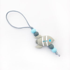 Peut inclure: Un hochet pour bébé en silicone gris et bleu avec un motif de fusée. Le hochet est attaché à un cordon gris.