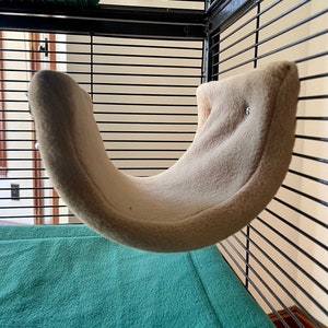 May include: A beige hammock-shaped bed for small pets, hanging inside a black wire cage. The bed is made of soft, plush fabric and has a curved shape.